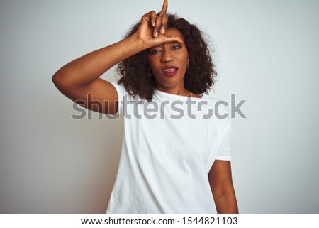 Young african american woman wearing t-shirt standing over isolated white background making fun of people with fingers on forehead doing loser gesture mocking and insulting.