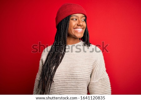 Young african american woman wearing winter sweater and wool hat over red isolated background looking away to side with smile on face, natural expression. Laughing confident.