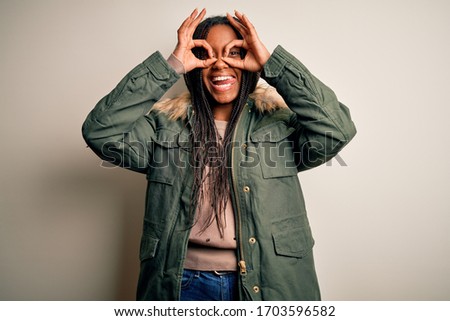 Young african american woman wearing winter parka coat over isolated background doing ok gesture like binoculars sticking tongue out, eyes looking through fingers. Crazy expression.