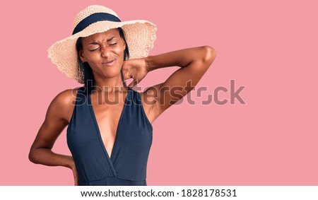 Young african american woman wearing swimsuit and summer hat suffering of neck ache injury, touching neck with hand, muscular pain 