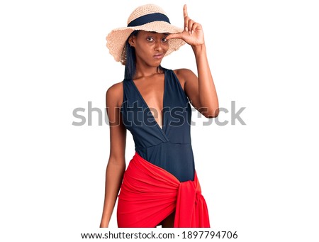 Young african american woman wearing swimsuit and summer hat making fun of people with fingers on forehead doing loser gesture mocking and insulting. 