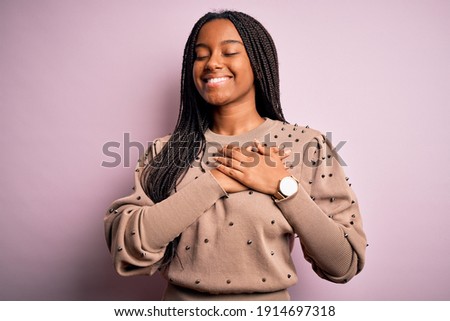 Young african american woman wearing fashion sweater over pink isolated background smiling with hands on chest with closed eyes and grateful gesture on face. Health concept.