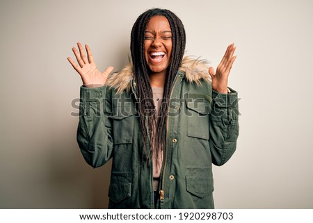 Young african american woman wearing winter parka coat over isolated background celebrating mad and crazy for success with arms raised and closed eyes screaming excited. Winner concept