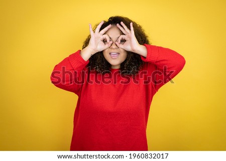 Young african american woman wearing red sweater over yellow background doing ok gesture shocked with smiling face, eye looking through fingers