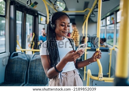 Young African american woman wearing earphones and listening to music on a smartphone while standing alone on a bus. Traveling to work and enjoying a bus ride. Portrait of a beautiful black woman.