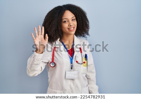 Young african american woman wearing doctor uniform and stethoscope waiving saying hello happy and smiling, friendly welcome gesture 