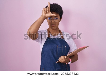 Young african american woman wearing professional waitress apron holding clipboard making fun of people with fingers on forehead doing loser gesture mocking and insulting. 
