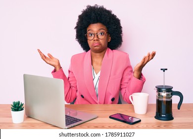 Young African American Woman Working At Desk Using Computer Laptop Clueless And Confused With Open Arms, No Idea And Doubtful Face. 