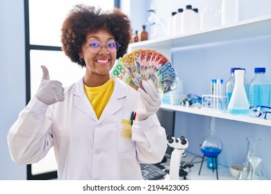 Young African American Woman Working At Scientist Laboratory Holding Money Smiling Happy And Positive, Thumb Up Doing Excellent And Approval Sign 