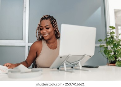 A young African American woman, working on documents at her desk, smiles while seated at her laptop. She is surrounded by a minimalist office setup with a potted plant adding greenery to the workspace - Powered by Shutterstock