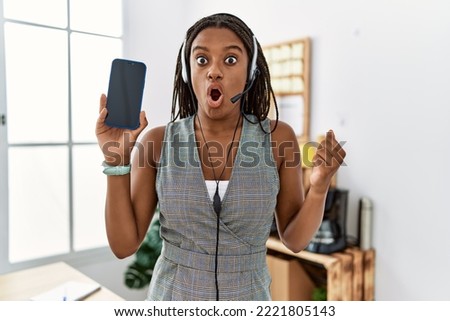 Young african american woman working at the office wearing operator headset holding smartphone scared and amazed with open mouth for surprise, disbelief face 