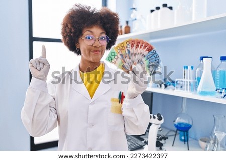 Young african american woman working at scientist laboratory holding money smiling with an idea or question pointing finger with happy face, number one 
