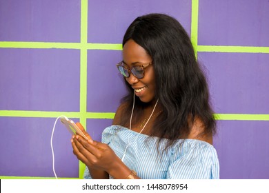 Young African American Women Playing Music Online With Her Mobile Phone Using Her Ear Piece To Listen.  Beautiful Girl Using Her Mobile Phone To Check Interesting Content Online