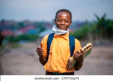 young African boy in uniform, with back pack and books in the hand-Black guy with face mask lowered at the chin with thumbs up sign-smiling  boy-school concept