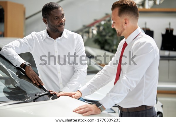 young african\
businessman ask questions about car presented in dealership,\
salesman explains and answer the questions, they sit on the hood\
and have friendly\
conversation