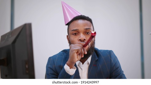 Young African Businessman Celebrating Birthday Alone In Office. Portrait Of Sad And Lonely Afro-american Employee Sitting At Workplace In Party Hat And Blowing Party Whistle