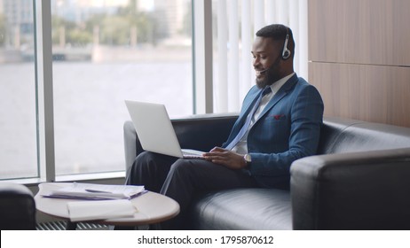 Young African Businessman In Headset Having Video Conference With Client On Laptop Sitting On Couch In Business Center Lobby. Afro Entrepreneur Relaxing On Sofa In Hall Having Video Call With Partner