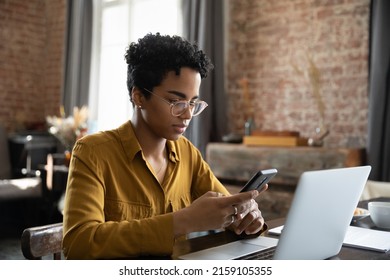 Young African businesswoman sit at workplace desk in front of laptop use smartphone, distracted from work on computer, using app chat online, synchronize files via application. Tech, workflow concept