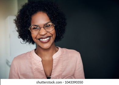 Young African businesswoman wearing glasses and laughing while standing alone in a modern office
