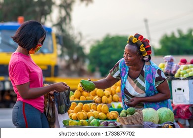 A young African buying fruits from the market and wearing face mask for protection - Receiving a purchased item from a local happy food vendor - Black millennial lifestyle in covid-19 pandemic season.
