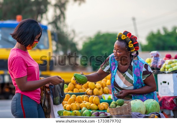 A young African buying fruits from the market and\
wearing face mask for protection - Receiving a purchased item from\
a local happy food vendor - Black millennial lifestyle in covid-19\
pandemic season.