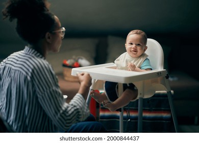 Young African, Caucasian Mother Feeding Her Mixed Race Son, He Is Cute And Having Fun, Loving, Tender Moment At Home . First Time Trying Solid Food, Love And Discovery