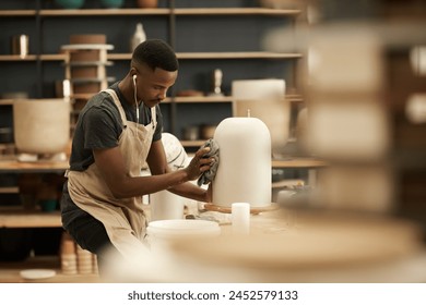 Young African ceramist wearing earphones putting the finishing touches on a pot while working in a pottery studio - Powered by Shutterstock