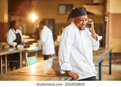 Young African chef leaning on a table in an industrial kitchen wearing chefs outfit on the phone placing orders at night  - Powered by Shutterstock