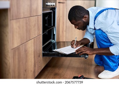 Young African Contractor Writing Information On Paper Document About Electric Oven Before Or After Repair, Side View On Confident Serious Guy Examining Inside Of Oven. Male In Blue Overalls, Workwear - Shutterstock ID 2067577739