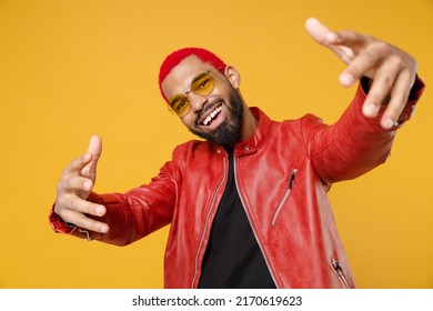 Young african cool rapper happy man 20s with funky trendy pink hairdo wear stylish red jacket glasses stretch hands to camera say yo yeah hi isolated on yellow orange color background studio portrait