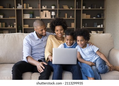 Young African couple with kids sit on sofa watch family movie online on laptop, use digital streaming services, buy goods, booking hotels for vacation, enjoy weekend at home with modern tech concept - Powered by Shutterstock