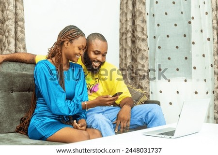 a young African couple seated comfortably on a sofa, radiating excitement as they gaze at the screen of a mobile phone held in their hands. With expressions of joy and engagement