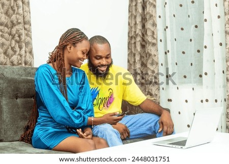 a young African couple seated comfortably on a sofa, radiating excitement as they gaze at the screen of a mobile phone held in their hands. With expressions of joy and engagement