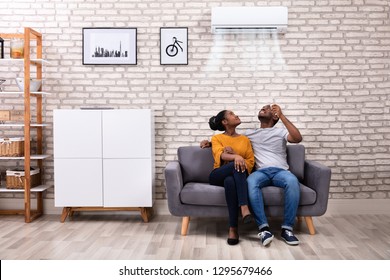 Young African Couple Sitting On Sofa Operating Air Conditioner At Home