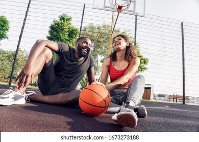 Young african descent couple sitting with ball on ground on basketball court outdoors laughing positive having fun bottom view