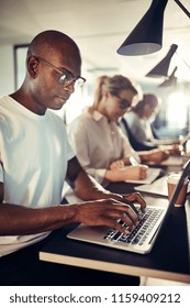 Young African Designer Focused On His Work While Sitting In A Row At A Table With Colleagues In A Modern Office