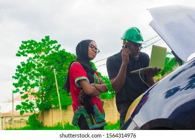 Young African engineer using his laptop to inspect a beautiful African lady’s car also making a phone call