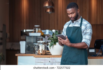 Young African entrepreneur in an apron standing in front of the counter of his trendy cafe using a digital tablet - Powered by Shutterstock