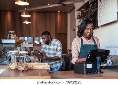 Young African Entrepreneur Working At The Checkout Counter Of A Trendy Cafe With A Coworker In The Background