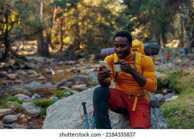 Young African Ethnicity Man Video Chatting Via Cellphone With Somebody And Drinking Hot Beverage As He Having A Hiking Walk In The Forest. Happy People, Network Technology, Or Traveling Concept