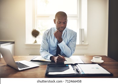 Young African Executive Sitting At His Desk In An Office Reading Documents And Working On A Laptop