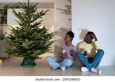 Young African Family Couple Resting After Decorating Christmas Tree Together At Home During Festive Season, Black Man And Woman Sits On Floor Near Firtree, Drink Coffee Or Tea In Cozy Xmas Atmosphere