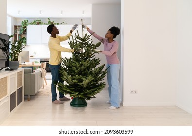 Young African Family Man And Woman Wearing Textile Gloves Straightening Branches Of Real Christmas Tree, Decorating House For Winter Holidays Together, Celebrating First Christmas As Married Couple