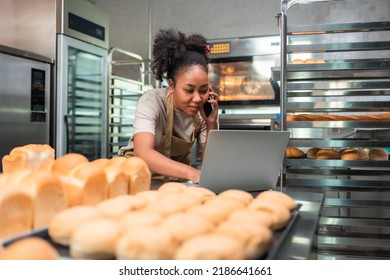 Young  African female in apron using laptop and talking to clients on the phone by workplace.Woman seller browsing online on cellphone. Business concept. Retail industry. - Powered by Shutterstock