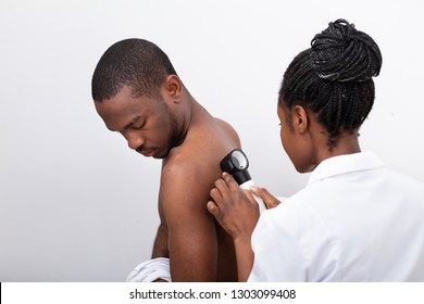 Young African Female Doctor Examining Pigmented Skin On Man's Back With Dermatoscope