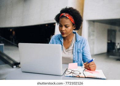 Young African female entrepreneur sitting at a table in a modern office building lobby working on a laptop and writing notes in her planner
