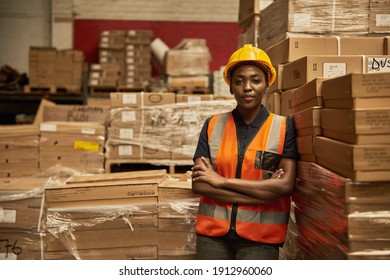 Young African female warehouse worker wearing a safety vest and helmet leaning with her arms crossed against a stack of boxes - Powered by Shutterstock