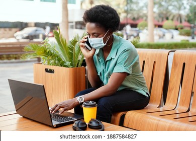 A young African girl in a medical face mask is talking on the phone and working with a laptop at a table in a cafe. Social distancing and work, work online, business online