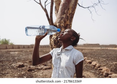 Young african girl in a parched field in front of a withered tree drinking water from a bottle; concept of lack of drinking water in equatorial regions
