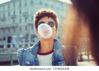 Young African Girl In Sunglasses With Curly Hair Puffs Bubble Of Chewing Gum In City Street. Brazilian Young Woman Inflates Pink Chewing Gum. Stylish Woman Making Bubblegum Of Chewing Gum.